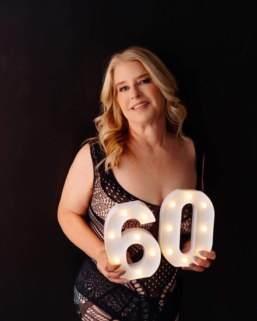 After: the same client in a black lace dress holding a lit-up 60 sign for her birthday