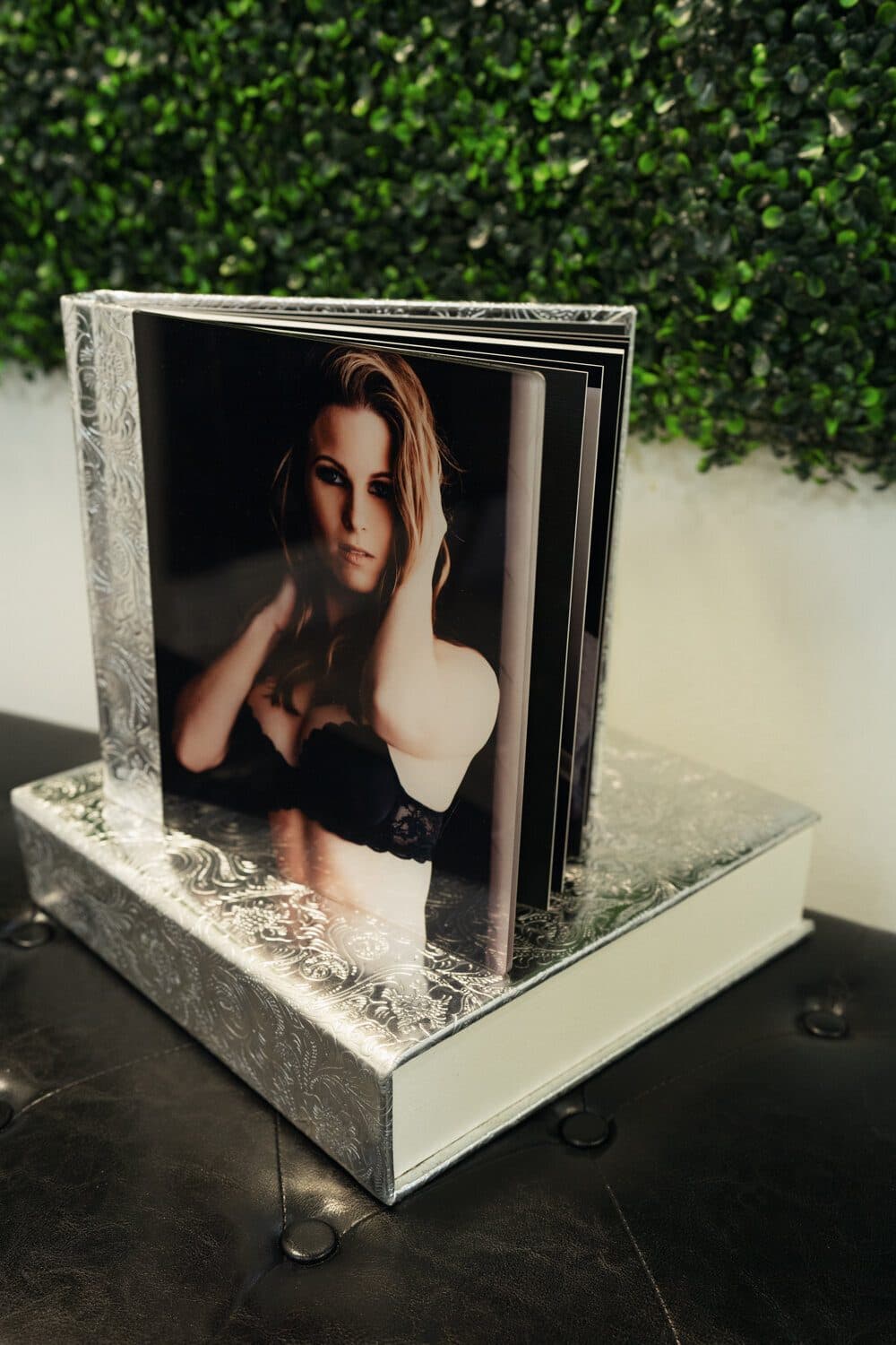 A silver embossed keepsake album stands open on its box, showing a portrait of a client in black lace, in front of a green hedge wall