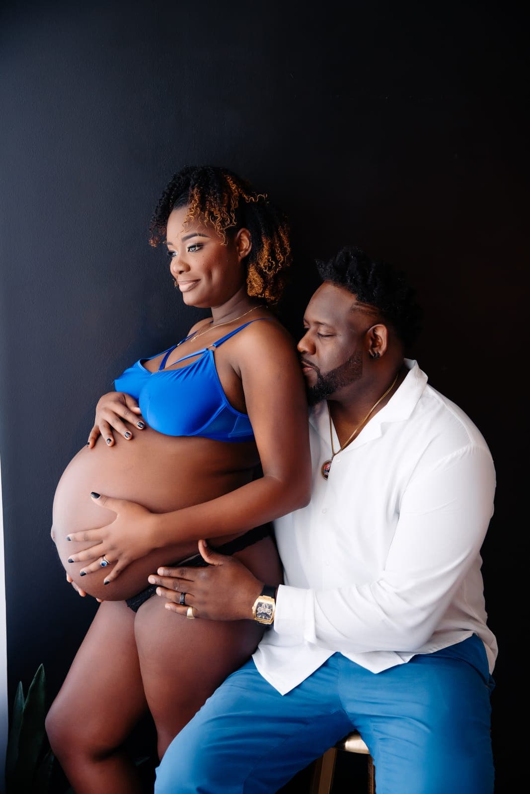 A pregnant client in a blue bra holds her belly while her partner in a white shirt sits beside her and kisses her shoulder
