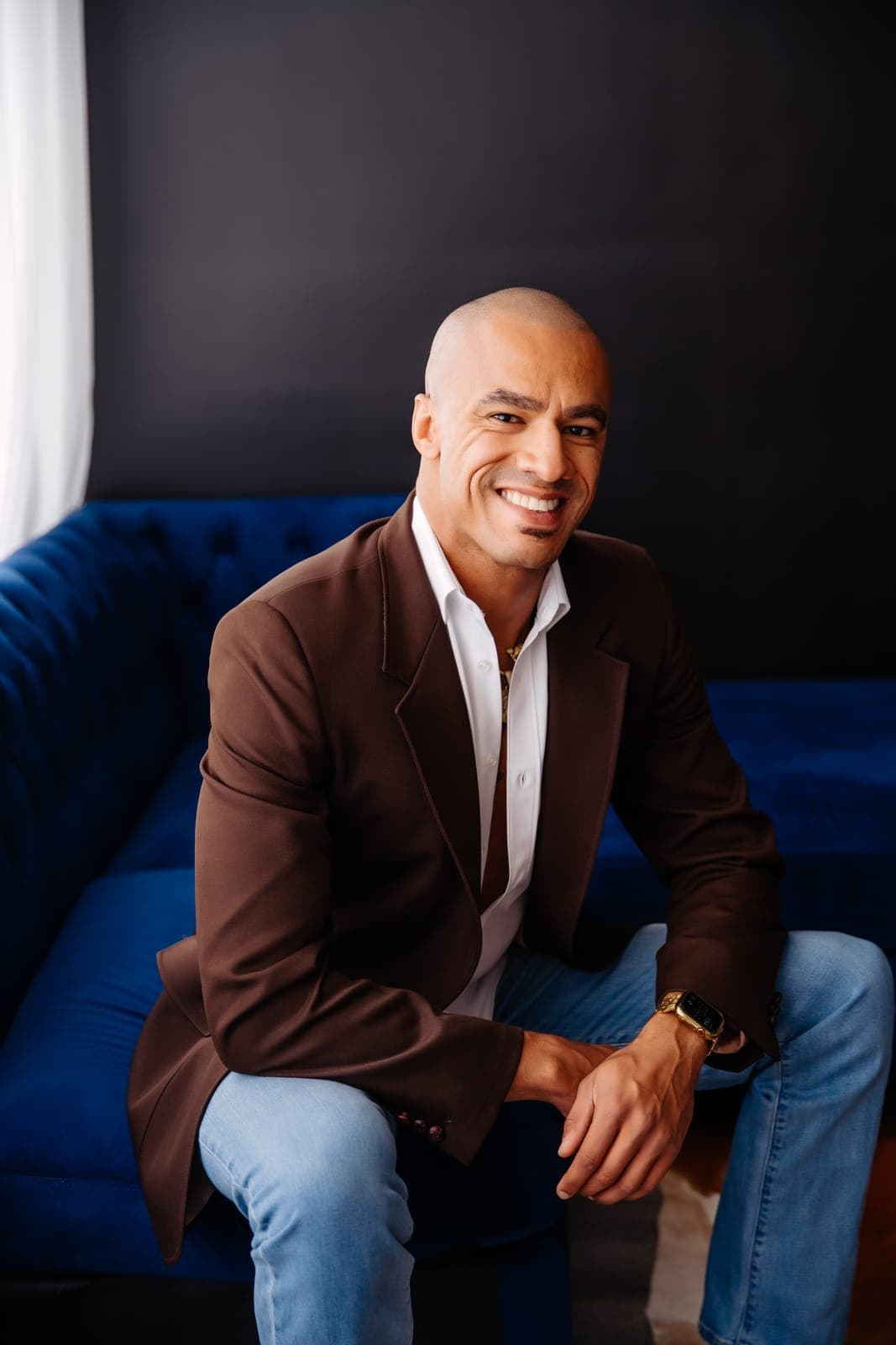 A smiling man in a brown blazer and jeans sits on the navy velvet couch