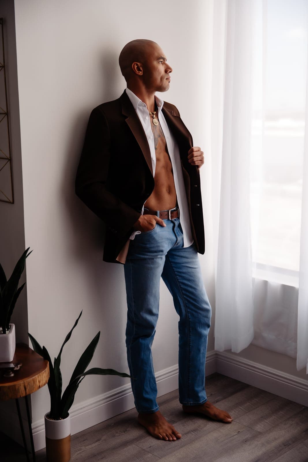 A man in a brown blazer and jeans stands barefoot by a bright window and looks out