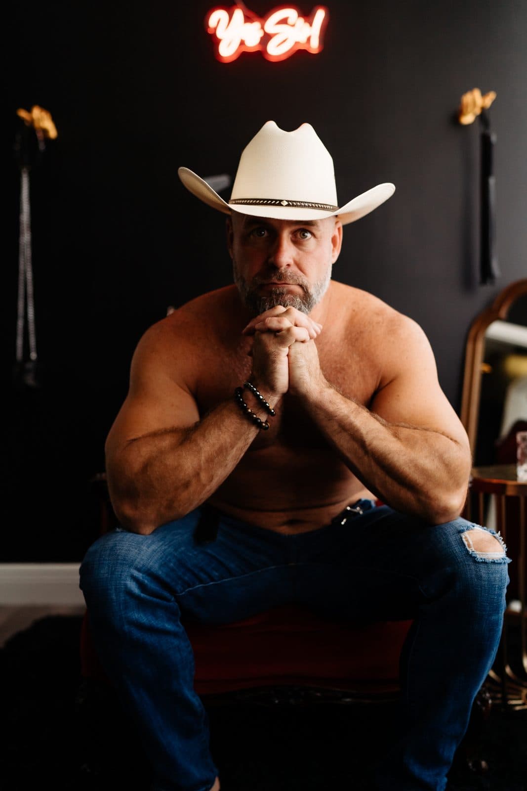 A man in a white cowboy hat and jeans, shirtless, under a neon sign