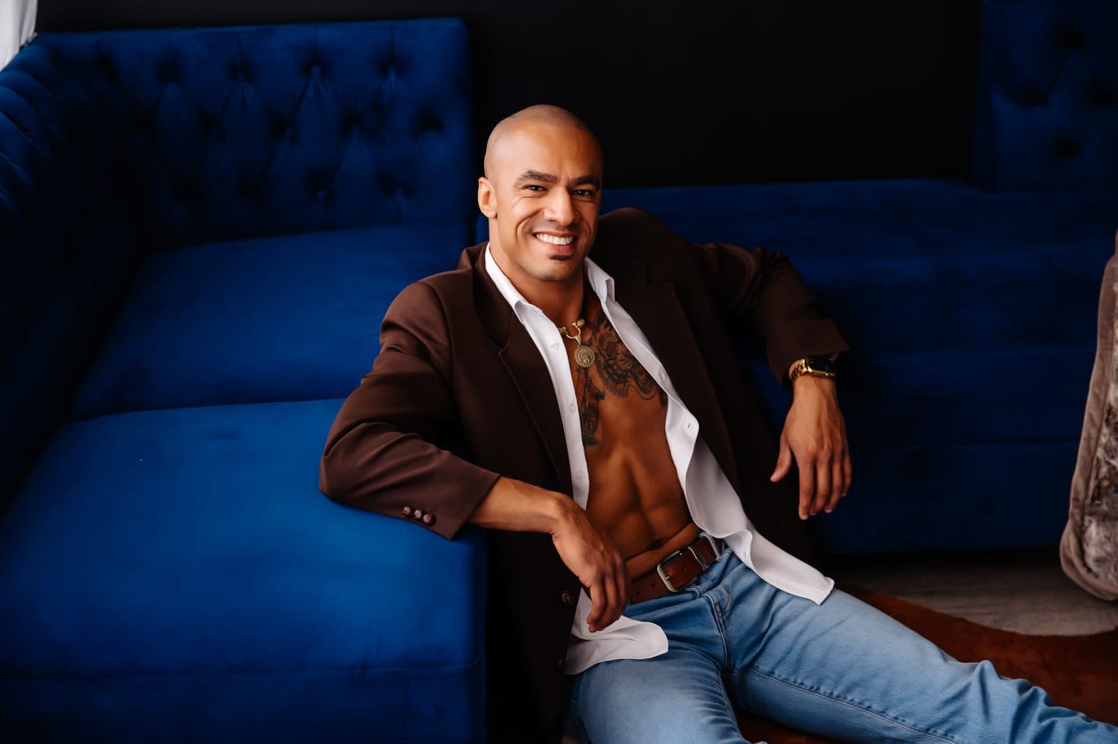 A smiling man in a brown blazer and jeans sits on the navy velvet couch