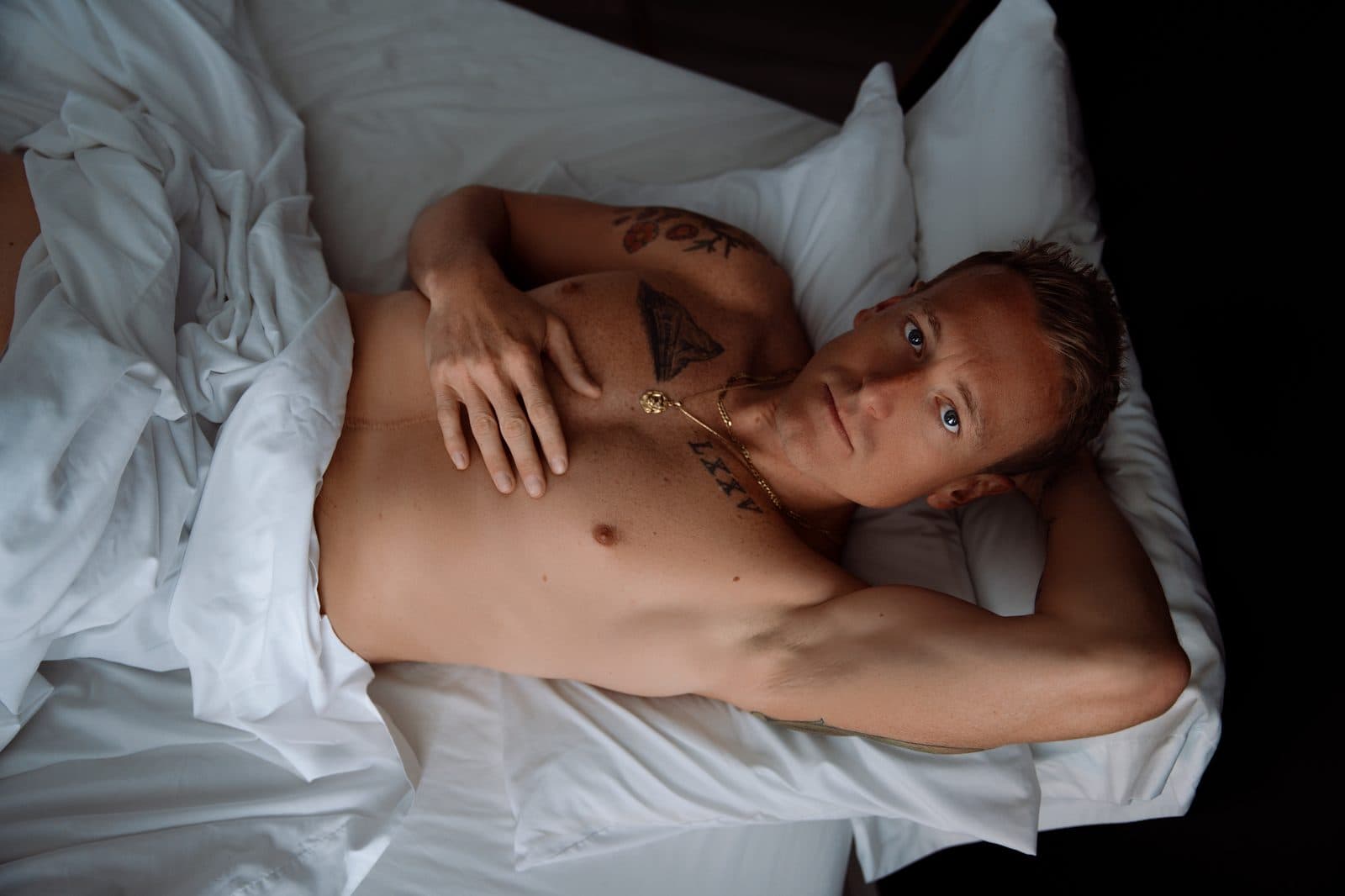 A man lying back on a white bed with a sheet over his hips, looking at the camera