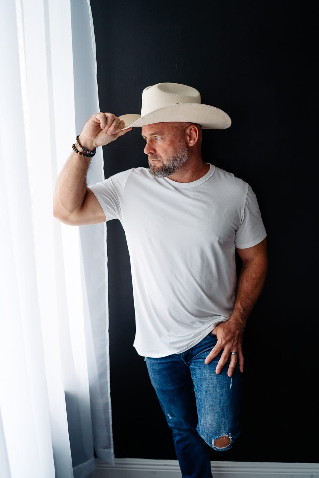 A man in a white cowboy hat and a white tee stands by a sheer window curtain