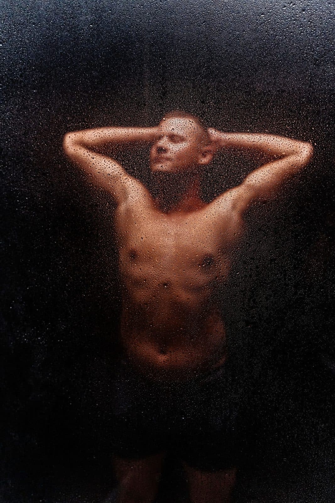 A man stands behind a water-beaded glass pane with his hands behind his head