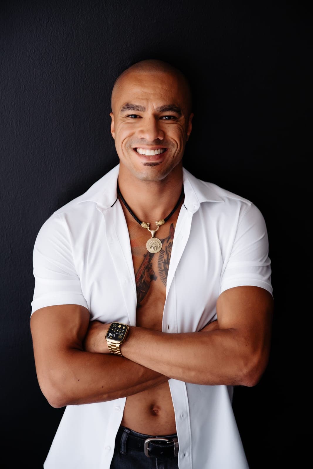 A smiling man with tattoos in an open white shirt crosses his arms against a black backdrop