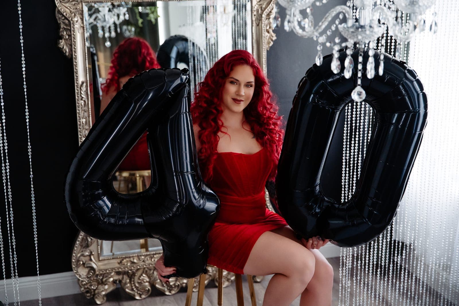A client with long red curls in a red dress sits between two big black 4 and 0 balloons in front of a silver mirror