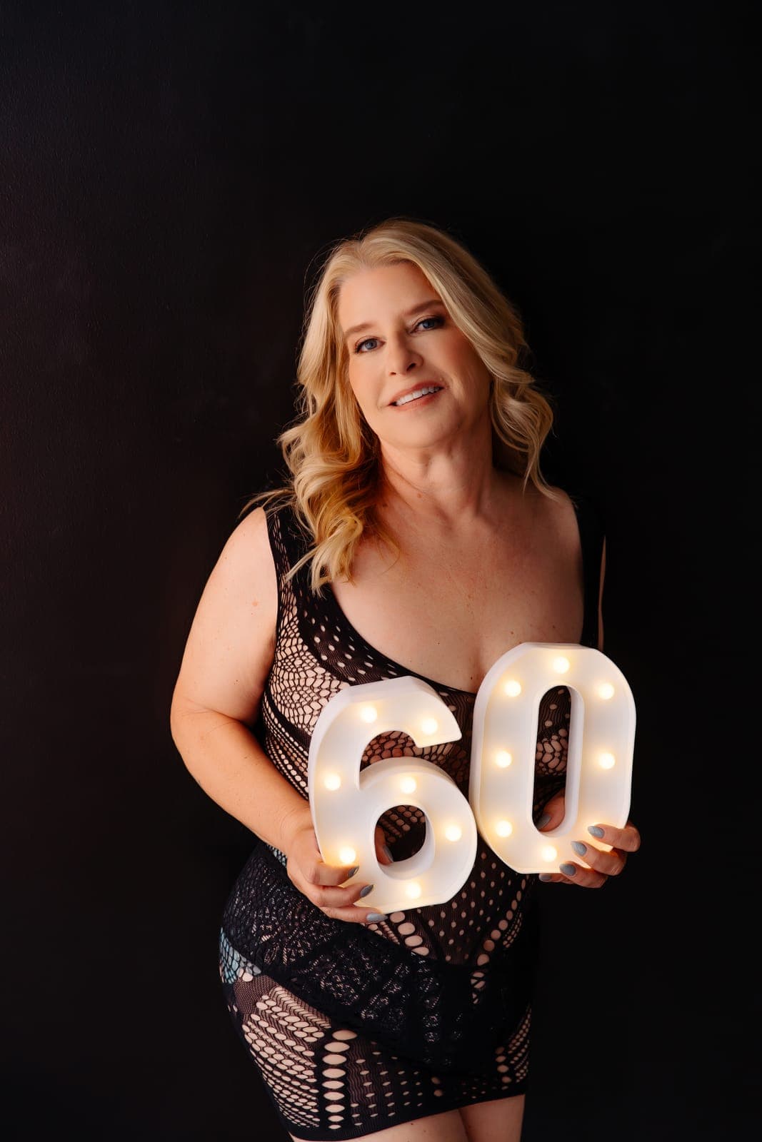 A smiling blonde client in a black lace dress holds a lit marquee number 60