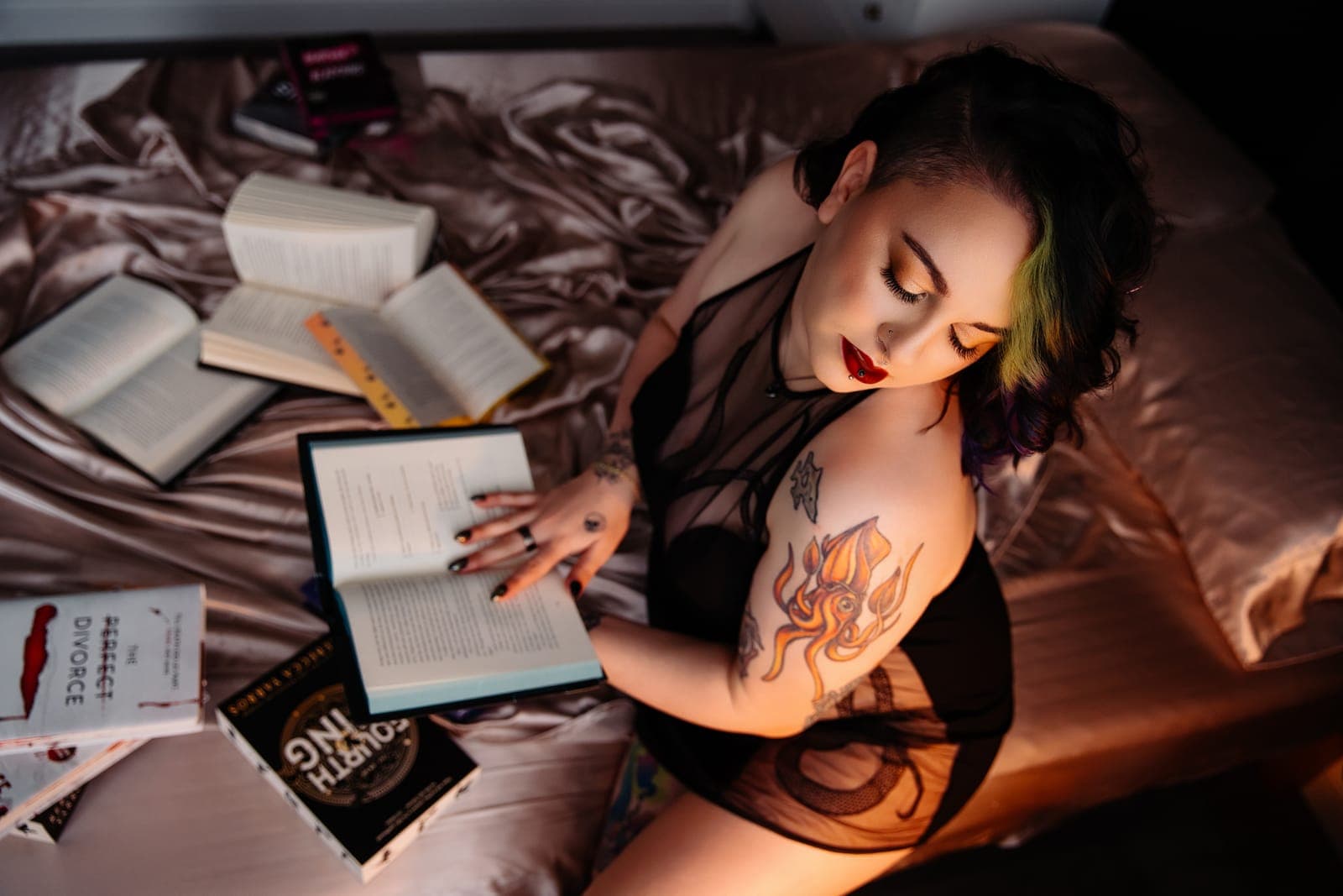 A client with tattoos and a green streak in her hair reads in bed, with open books spread over champagne satin sheets