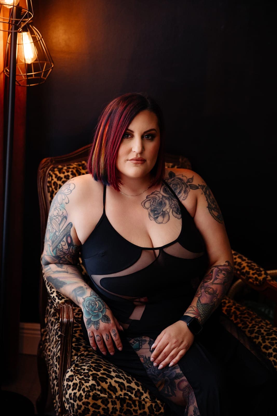 A client with tattooed arms in a black mesh dress sits in a leopard print chair beside a warm lamp