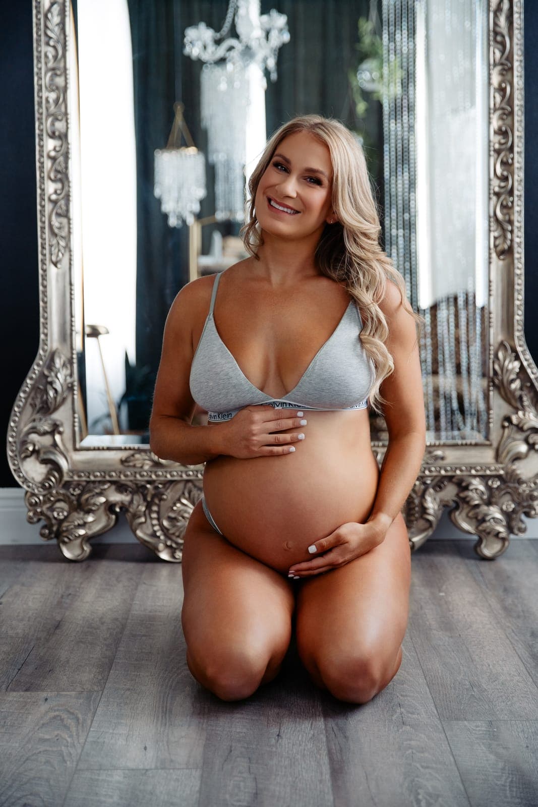 A smiling pregnant client in a gray bralette kneels in front of a tall silver mirror with her hands on her belly
