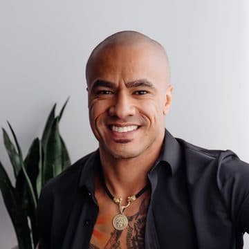 A smiling man in an open black shirt and jeans sitting on the navy velvet couch