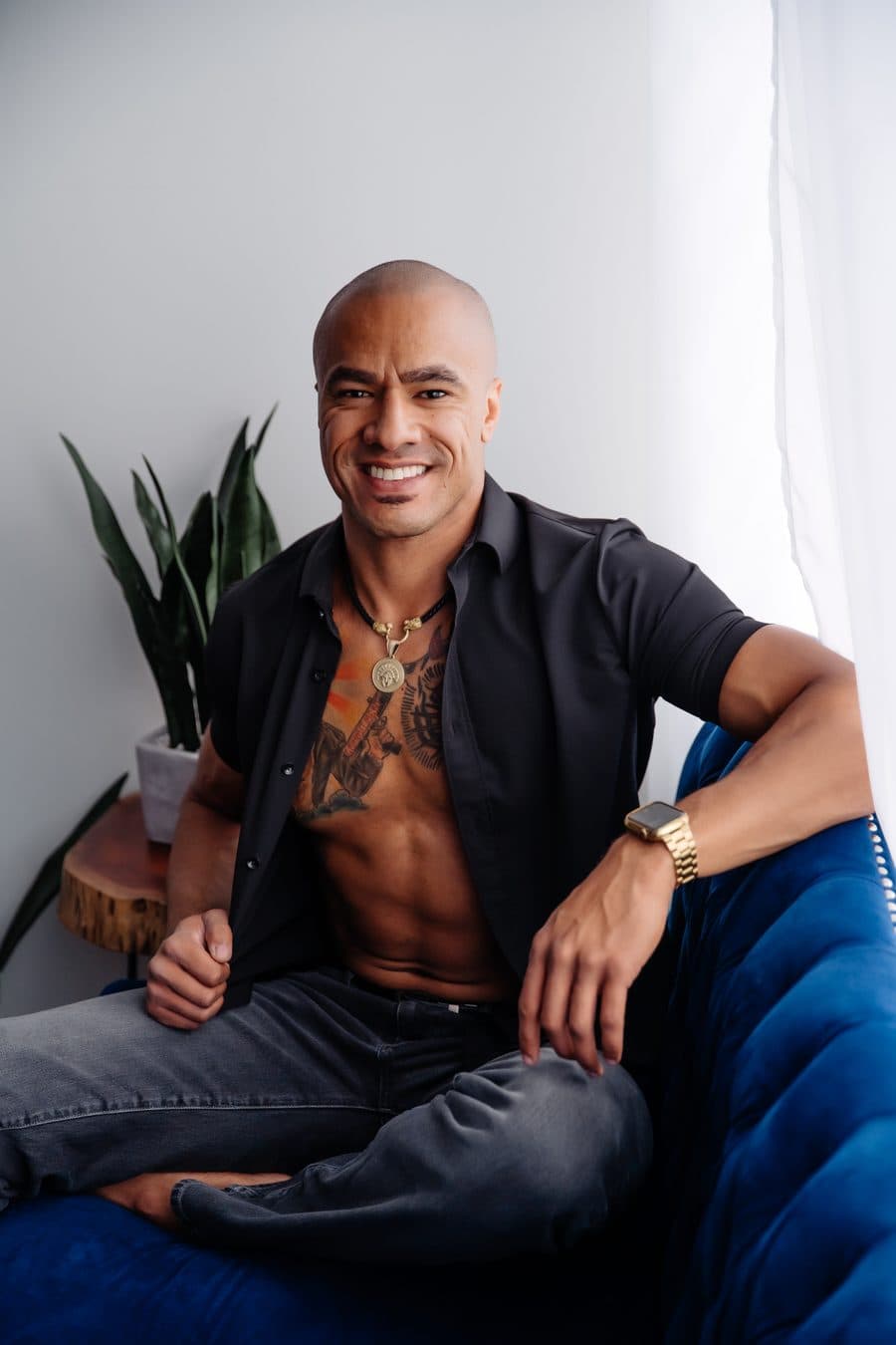 A smiling man in an open black shirt and jeans sitting on the navy velvet couch
